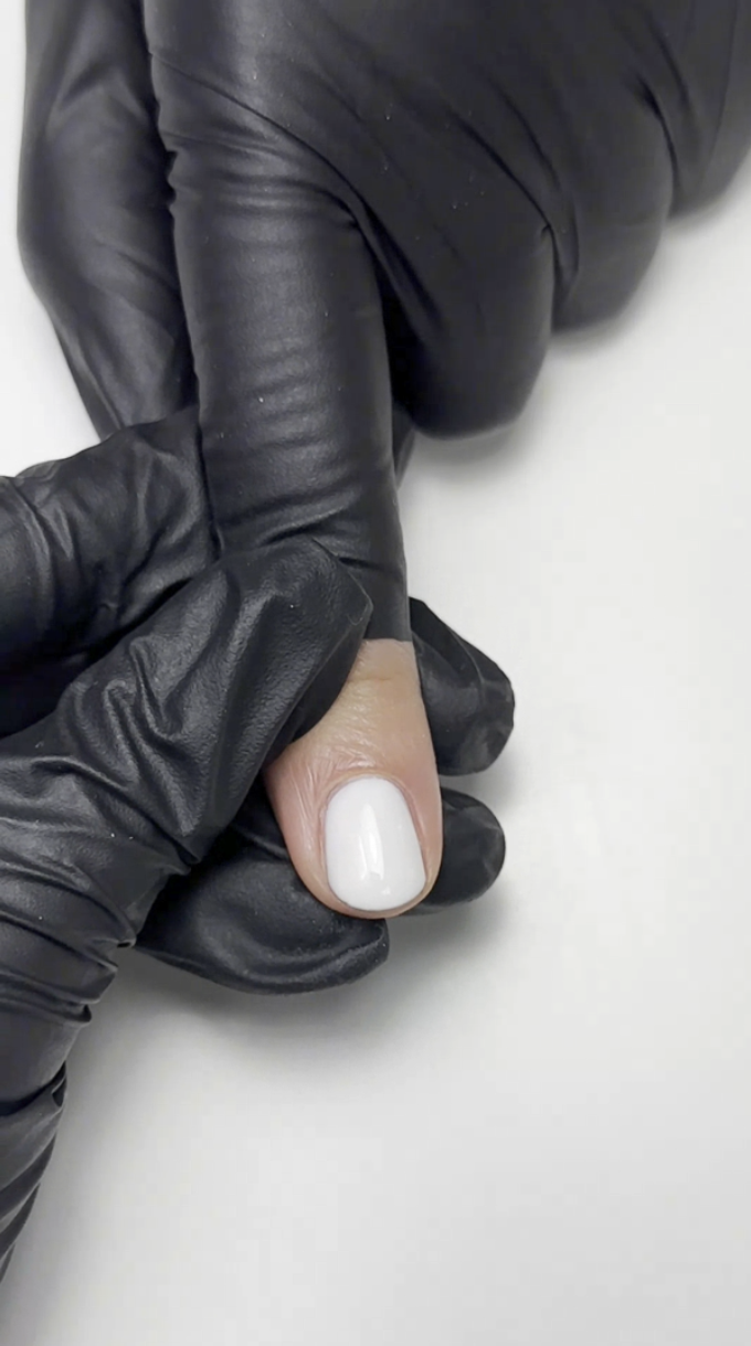  The image depicts the application stage in nail care, with a close-up of a person in black gloves applying a white nail polish to a fingernail, indicative of a precise manicure process.