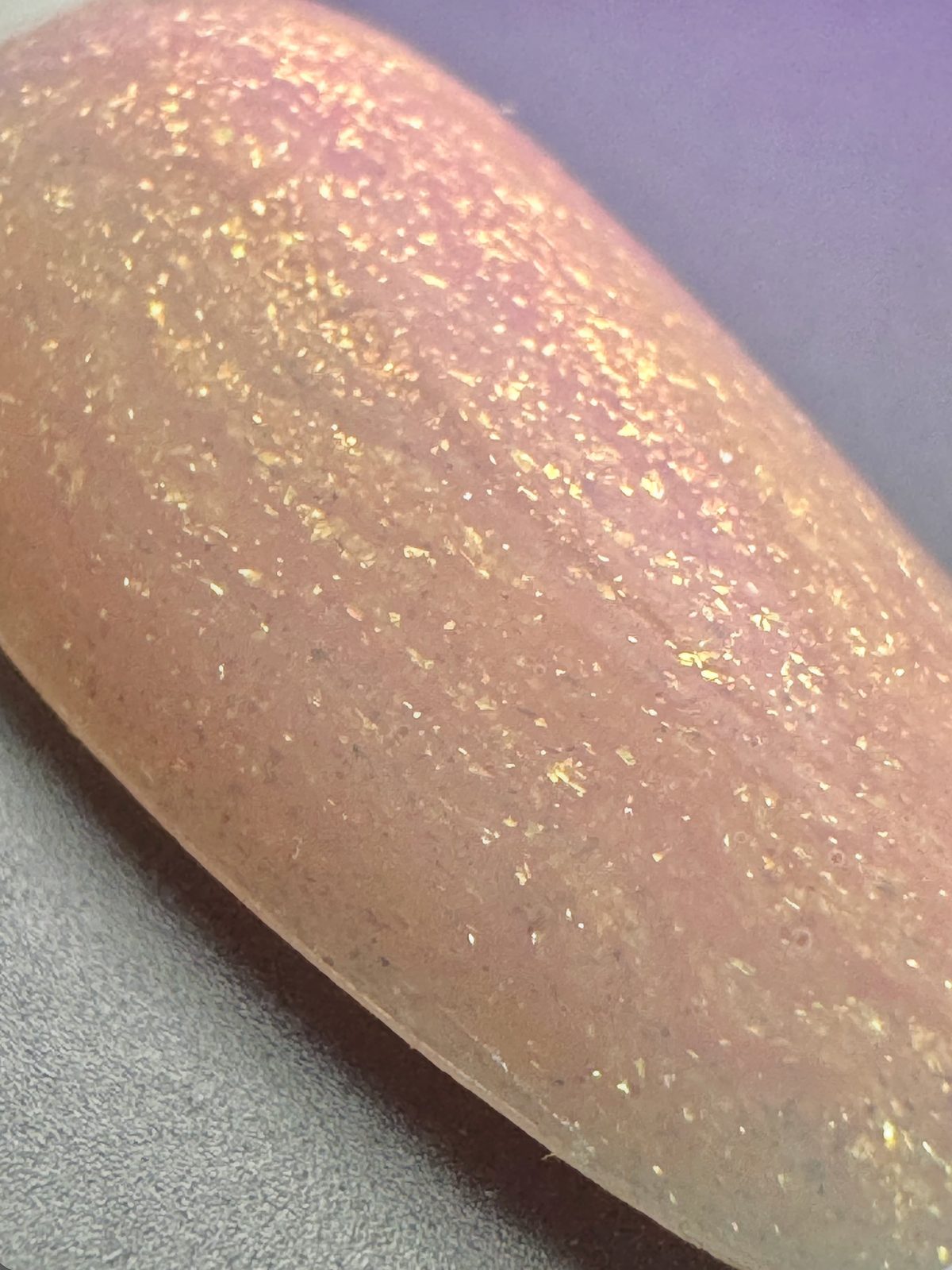 A close-up of a nail tip painted with a sheer, warm nude-pink base and heavily textured with shimmering gold glitter.