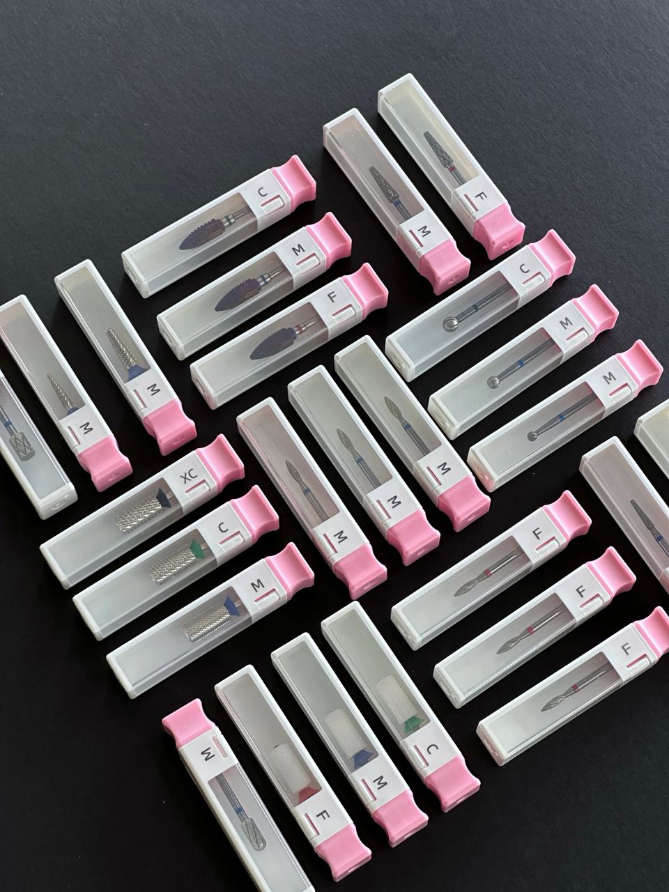  The image shows a collection of nail drill bits from MUSE, organized in a square pattern on a dark background, each piece encased in an individual clear container with a pink lid, labeled with different letters indicating the bit type.