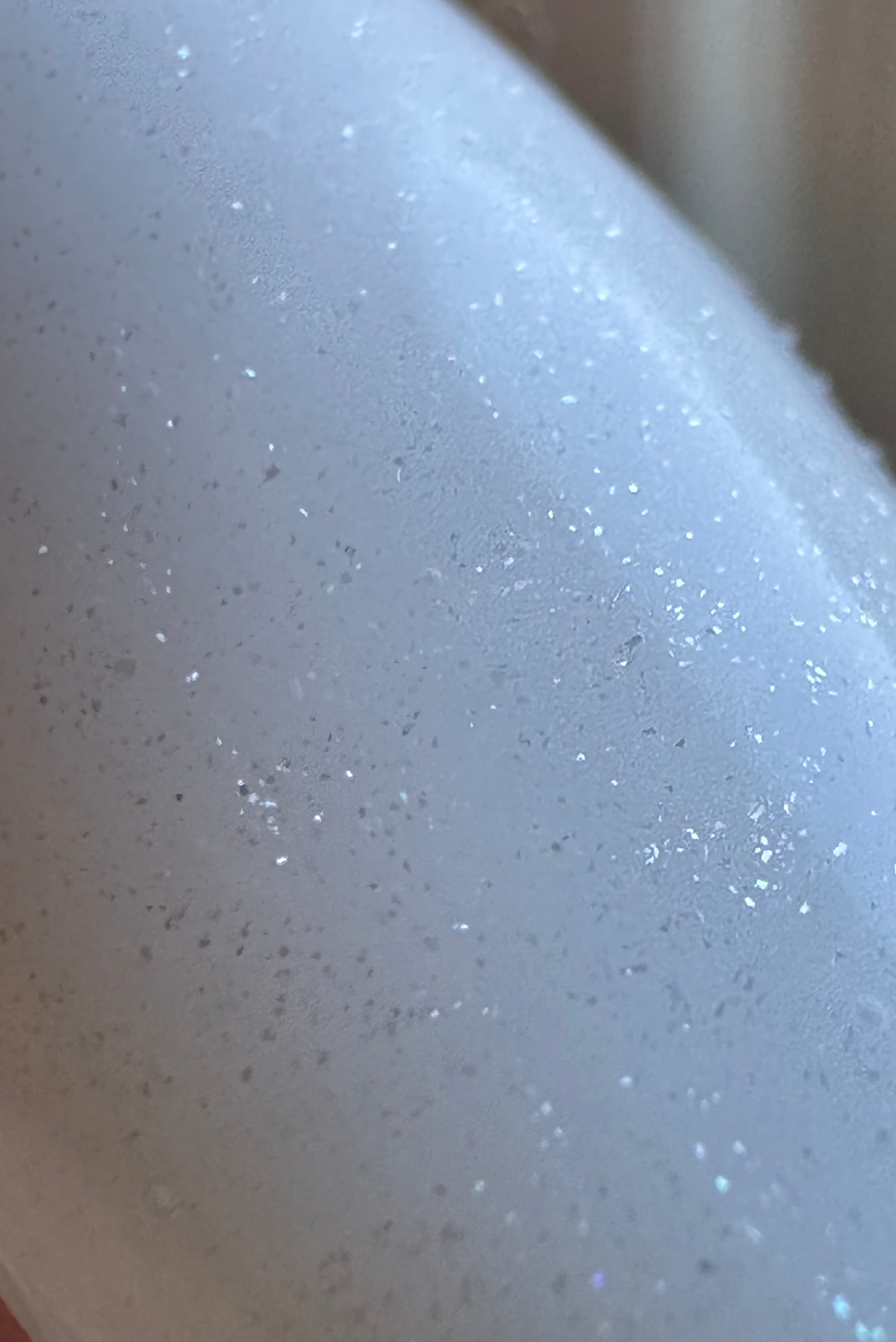 A close-up of a glossy white surface covered in fine silver glitter and small water droplets, with a soft warm-toned blurred background.