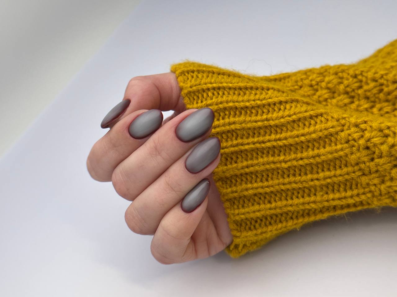 This image features a close-up of a hand with almond-shaped nails painted in a glossy grey color. The hand is elegantly posed, with the fingers slightly curled inward. In the background, there's a blush-colored cosmetic product with a golden cap, possibly a bottle of nail polish or skin care product, and a part of a bright yellow knitted garment or fabric, which adds a pop of color to the image. The focus on the hand against the soft background emphasizes the nail color and the sophisticated pose.