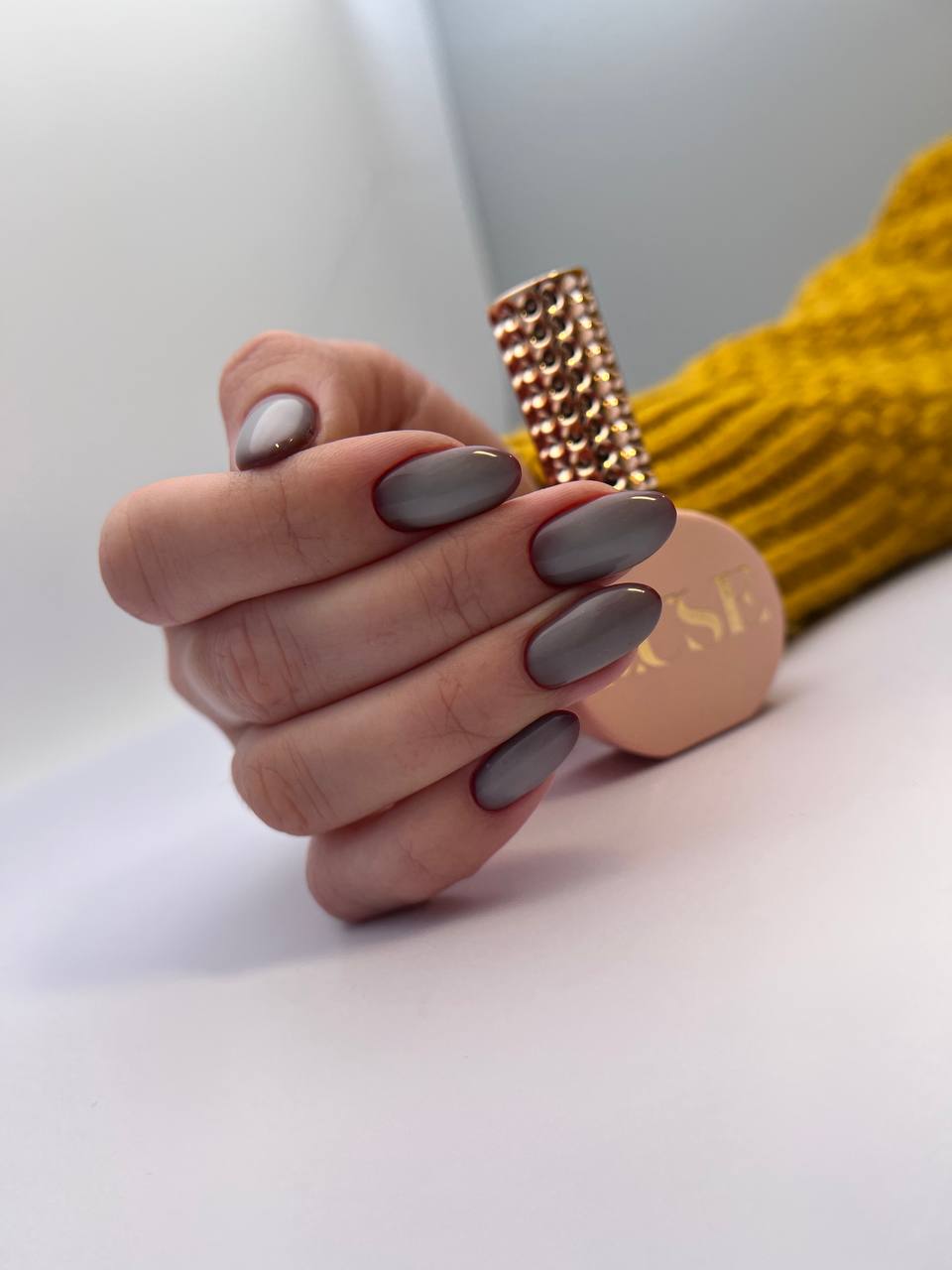 This image features a close-up of a hand with almond-shaped nails painted in a glossy grey color. The hand is elegantly posed, with the fingers slightly curled inward. In the background, there's a blush-colored cosmetic product with a golden cap, possibly a bottle of nail polish or skin care product, and a part of a bright yellow knitted garment or fabric, which adds a pop of color to the image. The focus on the hand against the soft background emphasizes the nail color and the sophisticated pose.