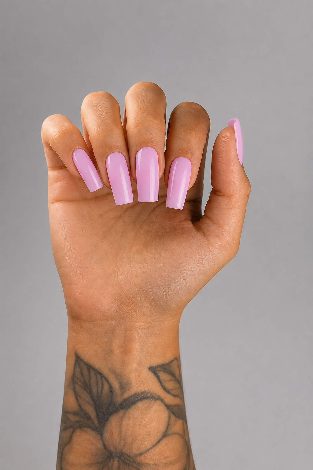 Close-up of long coffin-shaped nails finished in a glossy soft pastel pink gel color on a tattooed hand against a light grey background. Elegant professional nail design created with Muse Gel, featuring smooth full coverage and a high-shine finish.