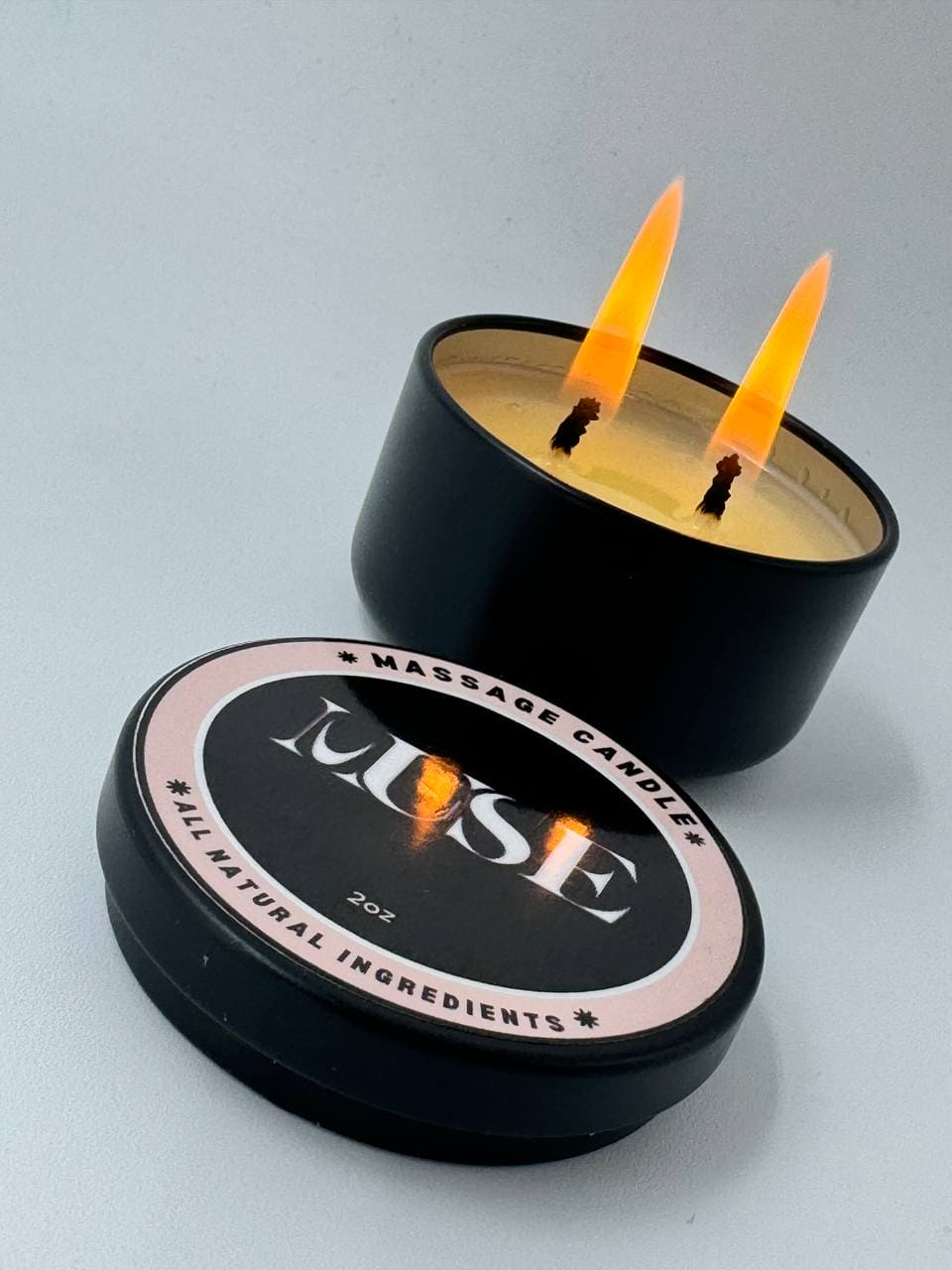  The image shows a lit MUSE massage candle with two bright flames, in a black container with the lid off to the side, highlighting the brand's focus on all-natural ingredients.